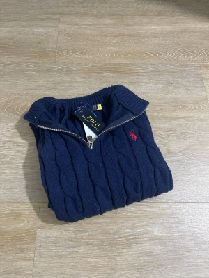 Mörkblå kabelstickad tröja Polo Ralph Lauren - Snygg mörkblå kabelstickad tröja från Polo Ralph Lauren med halv dragkedja och röd broderad logga på bröstet. Tröjan har hög krage och ribbade muddar. Perfekt för dig som gillar klassisk och stilren design.