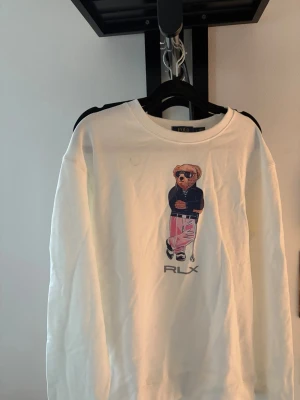 Vit sweatshirt med björntryck RLX - Vit sweatshirt från Polo Ralph Lauren med ett stort tryck av den ikoniska björnen i solglasögon, mörkblå jacka och rosa byxor. Rund halsringning och avslappnad passform. RLX-logga under motivet. Perfekt för en cool och avslappnad stil.