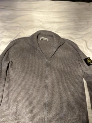 Grå ziptröja från Stone Island - Säljer en grå ribbstickad ziptröja från Stone Island med ikonisk patch på ärmen. Tröjan har hög krage, hel dragkedja och är tillverkad i ett mjukt stickat material. Perfekt för dig som gillar streetwear och snygga detaljer.