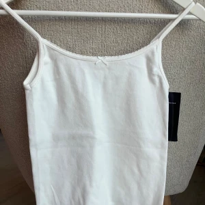 Vit linnetopp med rosett Brandy Melville - Söt vit linnetopp med tunna axelband och liten rosett framtill. Toppen är ribbad och har en enkel, stilren design som passar perfekt till många olika outfits. Materialet känns mjukt och bekvämt mot huden.