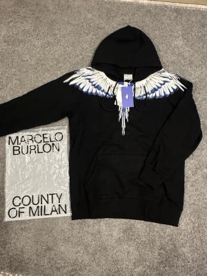Svart hoodie Marcelo Burlon vingar - Säljer en svart hoodie från Marcelo Burlon County of Milan med ikoniskt vingtryck i vitt och blått över axlar och bröst. Hoodien har huva med dragsko och en klassisk känguruficka framtill. Materialet är mjukt och skönt bomullstyg. Perfekt för dig som gillar streetwear med unik design.