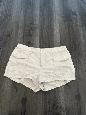 Beige shorts med fickor och knapp - Storlek M men mer som S/M. Snygga beige shorts med två stora fickor framtill och knappstängning. De har bälteshällor och en avslappnad passform, perfekta för varma dagar. Materialet känns lätt och luftigt, vilket gör dem sköna att bära hela sommaren.