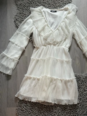 Vit volangklänning med v-ringning studentklänning - Supersöt vit kortklänning från GinaTricot med volanger längs v-ringningen och ärmarna. Klänningen har plisserat tyg, långa ärmar och markerad midja. Perfekt studentklänning eller sommaren för alla tillfällen 