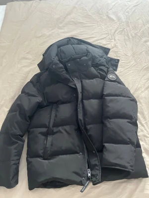 Svart dunjacka från Canada Goose (BLACK LABEL) - Säljer en svart dunjacka från Canada Goose med avtagbar huva och patch-logga på ärmen. Jackan har en klassisk puffer-modell och medföljer en pälskant till huvan. Perfekt för kalla vinterdagar och riktigt snygg streetstil.