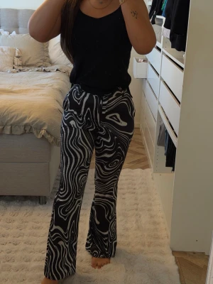 Svarta byxor med vit abstrakt mönster - Säljer ett par svarta byxor med vitt abstrakt mönster. Byxorna har en rak passform och är midwaist. Materialet känns mjukt och stretchigt, perfekt för dig som vill ha något bekvämt men ändå snyggt. Matcha med en enkel topp för en cool look.