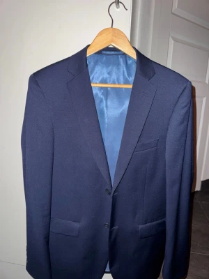 Mörkblå klassisk kavaj - Snygg mörkblå kavaj med två knappar framtill och klassiska fickor. Fodrad med matchande blått tyg på insidan och har en stilren, tidlös design. Perfekt för dig som vill ha en clean och elegant look.