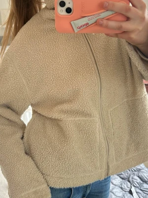 Beige teddyjacka med dragkedja - Mysig beige teddyjacka med huva och hel dragkedja framtill. Jackan har två stora fickor på sidorna och är gjord i fluffigt, mjukt teddy-material som håller dig varm. Perfekt för kalla dagar och enkel att matcha med jeans eller mjukisbyxor. Passar oversize på mig med S❤️