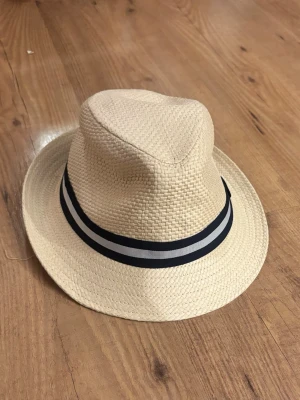 Beige stråhatt med randigt band - Snygg beige stråhatt med klassisk form och ett dekorativt svartvitt randigt band runt kullen. Tillverkad i flätat stråmaterial som ger en luftig känsla. Perfekt accessoar för soliga dagar och ger en chill vibe till din outfit.