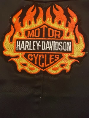 Svart Harley-Davidson jacka  med flames - Säljer en svart jacka från Harley-Davidson med ett stort broderat flames-märke på ryggen i orange, gult och vitt. Västen har klassisk bikerstil och är perfekt för dig som vill sticka ut på hojen. Grym detalj för MC-entusiasten! Jackan är nästan helt ny och har bara använts 2 gånger. Passar storlek S! 