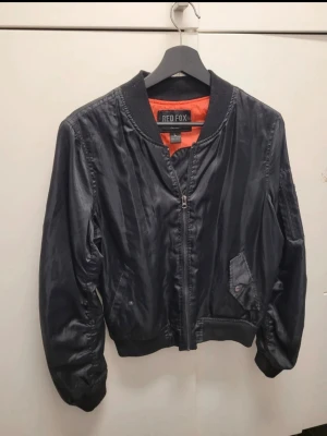Svart bomberjacka från Red Fox st S-L - Snygg svart bomberjacka från Red Fox med orange innerfoder. Jackan har klassisk ribbad krage, muddar och midja, samt dragkedja framtill. Kan passa en storlek S-L beroende på hur du vill att den ska sitta. :)