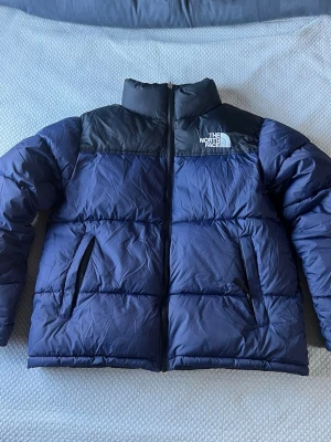 Blå och svart pufferjacka The North Face - Säljer en blå och svart pufferjacka från The North Face med hög krage och dragkedja framtill. Jackan har två sidofickor med dragkedja och klassisk logga broderad på bröstet. Perfekt för kalla vinterdagar och riktigt snygg streetstil.