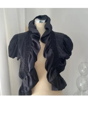 Svart volangblus från Sisters Point - Stilren svart blus från Sisters Point med korta puffärmar och volangkant framtill. Blusen har smockdetaljer längs sidorna och är tillverkad i ett glansigt, mjukt material som ger en lyxig känsla. Perfekt för att lyfta vilken outfit som helst.