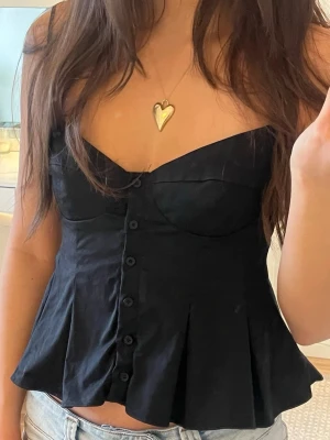 Svart korsettopp från Zara peplum - Säljer en svart korsettopp från Zara med knappar framtill och figurnära passform. Toppen har en snygg v-ringning och lätt utsvängd nederdel. Perfekt att styla med jeans eller kjol för en trendig look.