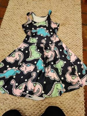 Svart klänning med dinosauriemönster - Söt svart klänning med färgglada dinosaurier i rosa, blått och grönt samt vita prickar. Klänningen har ärmlös design och rund halsringning. Perfekt för dig som vill sticka ut med ett lekfullt mönster. Väldigt stretchig 