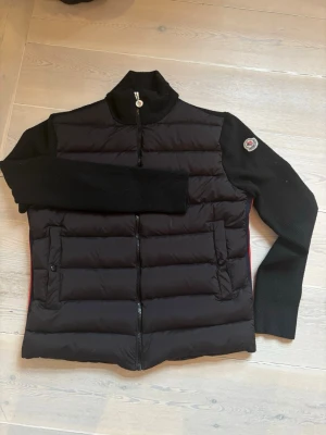 Svart jacka från Moncle - Svart jacka Moncle med stickade ärmar och hög krage. Framsidan har quiltad i dun och två fickor med knappar. Baksidan har stickat tyg och röda detaljer med -logga längs sidorna. Dragkedja framtill och märke på ärmen.