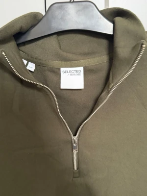 Olivgrön halfzip hoodie Selected Homme - Snygg olivgrön hoodie från Selected Homme med halv dragkedja framtill. Tillverkad i mjukt bomullsmaterial och har långa ärmar samt ribbade muddar. Perfekt för en avslappnad och trendig look.
