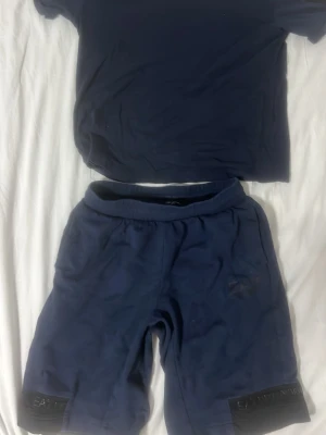 Mörkblå set från EA7 Emporio Armani - Säljer ett par mörkblå shorts o t shirt set från EA7 Emporio Armani med elastisk midja och logga på bensluten. Shortsen har en avslappnad passform och är tillverkade i mjukt bomullsmaterial, perfekta för sommardagar eller träning.