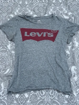 Grå Levi's t-shirt med röd logga - Säljer en klassisk grå t-shirt från Levi's med den ikoniska röda loggan tryckt på bröstet. T-shirten har korta ärmar och rund halsringning. Perfekt basplagg för en avslappnad stil.