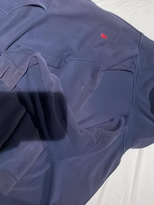 Blå hoodie från Polo Ralph Lauren - Mörkblå hoodie från Polo Ralph Lauren med dragkedja och klassisk röd logga broderad på bröstet. Tillverkad i mjukt material som känns skönt mot huden. Perfekt för en avslappnad och stilren look till sommaren.