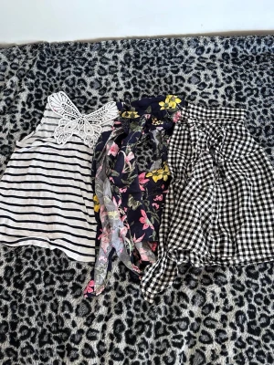 Tre somriga toppar i olika mönster - Säljer tre snygga toppar: en vit och svart randig linnetopp med spetsdetalj, en blommig omlottopp i mörkblått med rosa och gula blommor samt en svartvitrutig knytblus. Perfekta för varma dagar och att mixa och matcha!