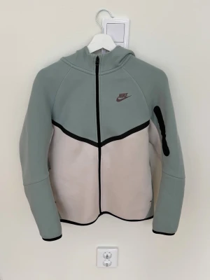 Nike hoodie i grått och vitt - Nike hoodie med dragkedja i en snygg ljusgrå och vit färgkombination. Hoodien har huva, svarta detaljer och Nike-logga på bröstet. Tillverkad i mjukt och stretchigt material, perfekt för en sportig och avslappnad stil.