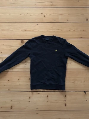 Svart sweatshirt från Lyle & Scott - Klassisk svart sweatshirt från Lyle & Scott med rund halsringning och det ikoniska gula örnlogot på bröstet. Tröjan har långa ärmar och ribbade muddar vid ärmslut och nederkant. Perfekt för en clean och stilren look.