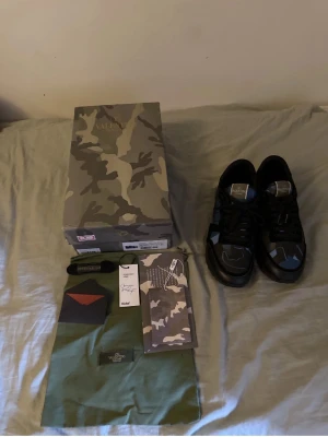 Valentino Garavani Camo Sneakers - Snygga sneakers från Valentino Garavani med svart bas och camo-detaljer i blått och grått. Skorna har klassisk låg profil, rund tå och svarta skosnören. Kommer med originalkartong, extra skosnören och dustbag. Perfekt för dig som gillar exklusiv streetwear.