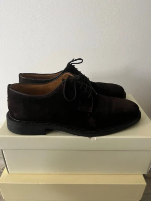 Mörkbruna mocka loafers - Säljer ett par stilrena mörkbruna loafers i mocka med klassisk rund tå och låg klack. Skorna har snörning framtill och en diskret söm längs sulan. Insidan är fodrad i ljust läder för extra komfort. Perfekta för dig som gillar en tidlös och clean look.