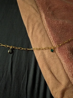 Guldhalsband med grönt hänge - Snyggt halsband i guldfärgad metall med två hängen, ett genomskinligt och ett smaragdgrönt. Kedjan har större, avlånga länkar som ger ett trendigt intryck. Perfekt för dig som gillar stilrena detaljer med en twist.