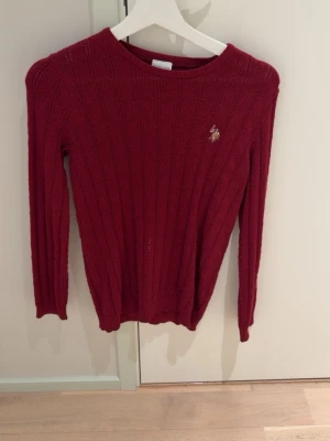 Vinröd stickad tröja U.S. Polo Assn. - Snygg vinröd stickad tröja från U.S. Polo Assn. med diskret logga broderad på bröstet. Tröjan har rund halsringning och ribbade muddar vid ärmslut och nederkant. Perfekt till höst och vinter för en stilren look.