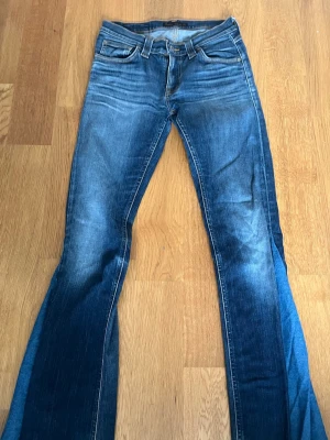 Blå bootcut jeans från Nudie Jeans - Säljer ett par blå bootcut jeans från Nudie Jeans med klassisk femficksmodell och snygga slitningar. Jeansen har kontrastsömmar, vågig brodyr på bakfickorna och råa, fransiga benslut. Perfekta för en avslappnad och trendig look.