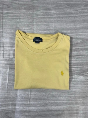Gul t-shirt från Polo Ralph Lauren - Snygg gul t-shirt från Polo Ralph Lauren med klassisk rund hals och liten broderad logga på bröstet. Tillverkad i mjuk bomull som känns skön mot huden. Perfekt för en avslappnad och stilren look.