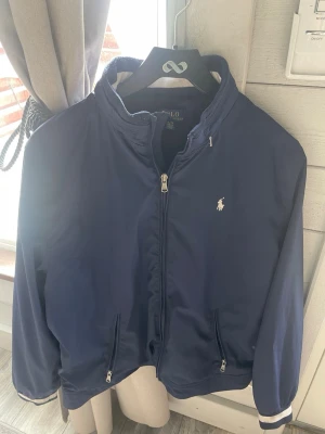 Marinblå vindjacka Polo Ralph Lauren - Snygg marinblå vindjacka från Polo Ralph Lauren med dragkedja framtill, två fickor med dragkedja och vit broderad logga på bröstet. Jackan har ribbade muddar med vit rand och står för en klassisk och sportig stil. Passar mig som storlek S jag är 170cm 65 kg men passar säkert något barn som XL små skador men funkar helt perfekt. 