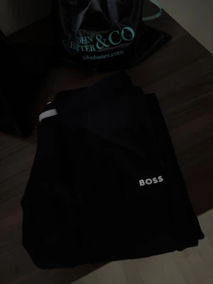 Svarta mjukisbyxor från Hugo BOSS - Säljer ett par svarta mjukisbyxor från BOSS med vit logga på benet. Byxorna har dragsko i midjan och smal passform. Perfekta för chill dagar eller när du vill ha en clean och enkel look. Materialet känns mjukt och bekvämt.