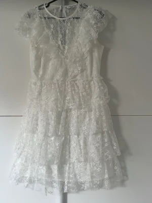 Vit spetsklänning med volanger - Superfin vit kortklänning i spets med volangdetaljer och v-ringning. Klänningen har korta ärmar och flera lager spets som ger en romantisk känsla. Perfekt för sommarens alla tillfällen. Ingen synlig branding. Öppen för prisförslag