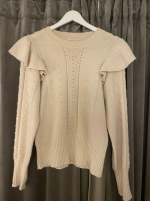 Beige stickad tröja med volang - Supersnygg beige stickad tröja från OBJECT med volangdetaljer på axlarna och strukturstickat mönster. Tröjan har rund halsringning, långa ärmar och breda ribbade muddar. Perfekt för dig som gillar en trendig och unik look.