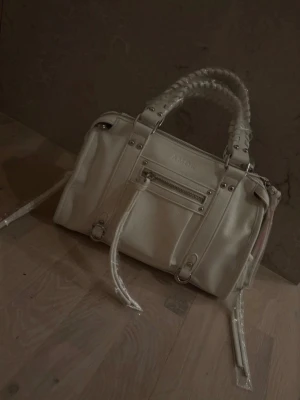 Vit handväska  - En jättefin vit väska från design by Amor. Väskan är helt ny och oöppnad med plast och lappar kvar. Jag säljer denna väska eftersom jag råkade köpa två väskor. Storleken på väskan är 30x14x20 cm. 