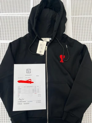 Svart hoodie med röd Ami-dekal - Svart hoodie från Ami med dragkedja framtill och huva med snören. På bröstet sitter en röd Ami-dekal i form av ett hjärta med bokstaven A. Hoodien har ribbade muddar och fickor på sidorna. Perfekt för en avslappnad och trendig look.