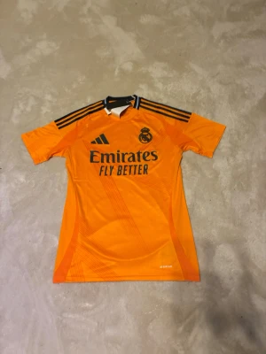 Orange Real Madrid fotbollströja Adidas - Snygg orange Real Madrid fotbollströja från Adidas med svarta detaljer och klubbmärke på bröstet. Tröjan har korta ärmar, rund hals och är tillverkad i lätt polyester som andas. Perfekt för dig som älskar fotboll och vill sticka ut på planen.