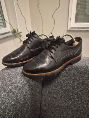 Jana Svarta loafers i äkta läder 38 - Säljer ett par klassiska svarta loafers i äkta läder med snörning och robust sula. Skorna har en stilren design med bruna detaljer längs sulkanten och passar perfekt till både jeans och kostym. Perfekt för dig som gillar tidlös och enkel stil.