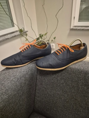 Zara mörkblå loafers med orange snören 44 - Säljer ett par stilrena mörkblå loafers med kontrasterande orange snören och ljus sula. Skorna har en slimmad, låg profil och är tillverkade i ett tygmaterial med innersula i brunt läderimitation. Perfekta för dig som vill sticka ut med färg på snörena.