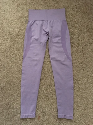 Ljuslila seamless leggings från NVGTN - Säljer ett par ljuslila seamless leggings från NVGTN med hög midja och diskreta paneldetaljer längs sidorna. Materialet är stretchigt och mjukt, perfekt för träning eller chill. Loggan sitter baktill i midjan. Supersnygg färg som sticker ut!