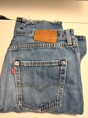 Levi's raka blå jeans med slitning - Klassiska blå jeans från Levi's med rak passform och knappgylf. Jeansen har en slitning på ena benet och den ikoniska röda Levi's-lappen på bakfickan. Tillverkade i robust denim med läderpatch bak i midjan. Perfekta för en avslappnad och trendig look.