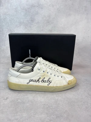 Saint Laurent vita sneakers 'yeah baby' - Vita sneakers från Saint Laurent med broderad text 'yeah baby' på sidan. Klassisk låg modell med vita skosnören, gummisula och detaljer i beige. Tillverkade i läder och textil för en clean och stilren look.