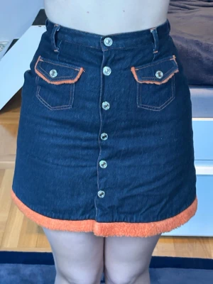 Mörkblå jeanskjol med orange detaljer - Säljer en mörkblå fodrad kort jeanskjol med orange fuskpälsdetalj längst ner och orange sömmar. Kjolen har framfickor med lock och knappar längs hela framsidan. Perfekt för dig som vill sticka ut med coola detaljer! 🧡💙