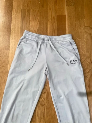 Gråa byxor från EA7 Armani - Snygga ljusgrå mjukisbyxor från EA7 Emporio Armani med dragsko i midjan och två sidofickor. Byxorna har en relaxed fit och EA7-logga tryckt på vänster ben. Perfekta för chill dagar eller när du vill ha en clean och stilren look. 