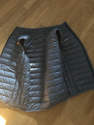 Grå dunväst från Ellesse - Snygg grå dunväst från Ellesse med quiltad design och diskret logga på bröstet. Västen har en klassisk passform och är lätt vadderad för extra värme. Perfekt att slänga över en hoodie eller tröja när det är kyligt ute.