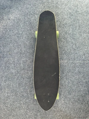 Svart longboard med limegröna hjul - Longboard med svart grepptejp på ovansidan och limegröna hjul. Brädan har en klassisk avlång form och är tillverkad i trä med flera lager. Perfekt för cruising och längre turer i stan eller parken.