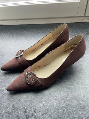 Bruna mockapumps med spänne - Bruna pumps i mockaimitation med spetsig tå och metallspänne framtill. Skorna har låg klack och är i storlek 38. Tidigare köpt på Sellpy men passade inte mig så säljer vidare de! Smutsiga på insidan men inget fel på utsidan av skorna!🥰