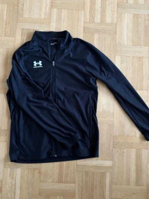 Svart träningsjacka från Under Armour - Snygg svart träningsjacka från Under Armour med dragkedja framtill och hög krage. Använd cirka 3-4 gånger kan gå ner i pris vid snabbt köp. Passar M lite liten i storlek. 